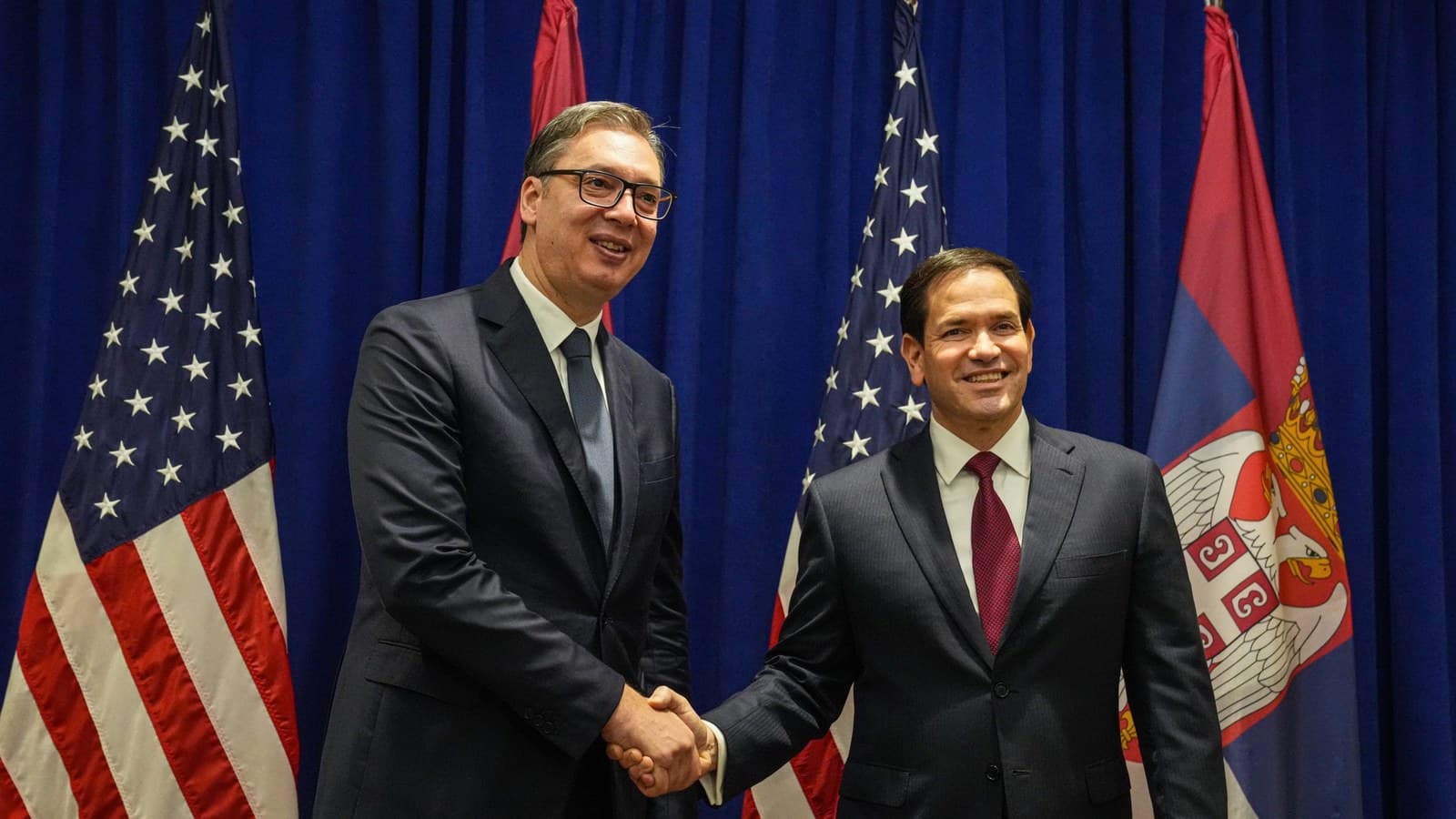 Aleksandar Vucic, in glasses and a dark suit, smiles and shakes hands with U.S. Secretary of State Marco Rubio in front of a blue curtain, U.S. flags and a Serbian flag.