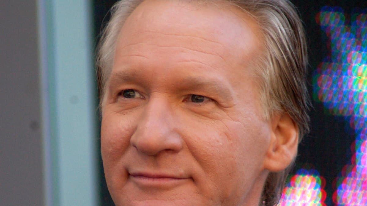 Primer plano del comediante y presentador Bill Maher, con una leve sonrisa y cabello canoso, al aire libre en Los Ángeles.
