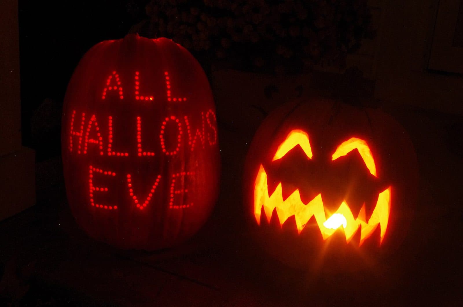Two lit jack-o'-lanterns at night, one carved with All Hallows Eve in dots and one with a jagged grin, 2012.