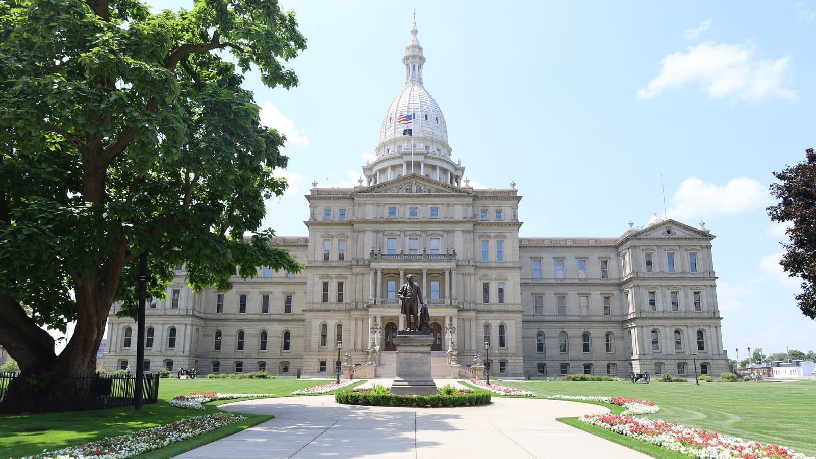 The domed Michigan State Capitol in Lansing on a sunny summer day, with a statue, flower beds and a walkway in front, in a July 2025 archive photo.