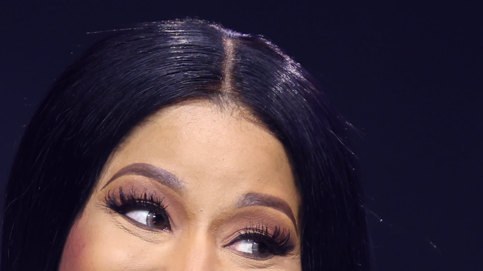 Nicki Minaj speaking into a microphone at AmericaFest in Phoenix, with long dark hair and a black top against a dark background.