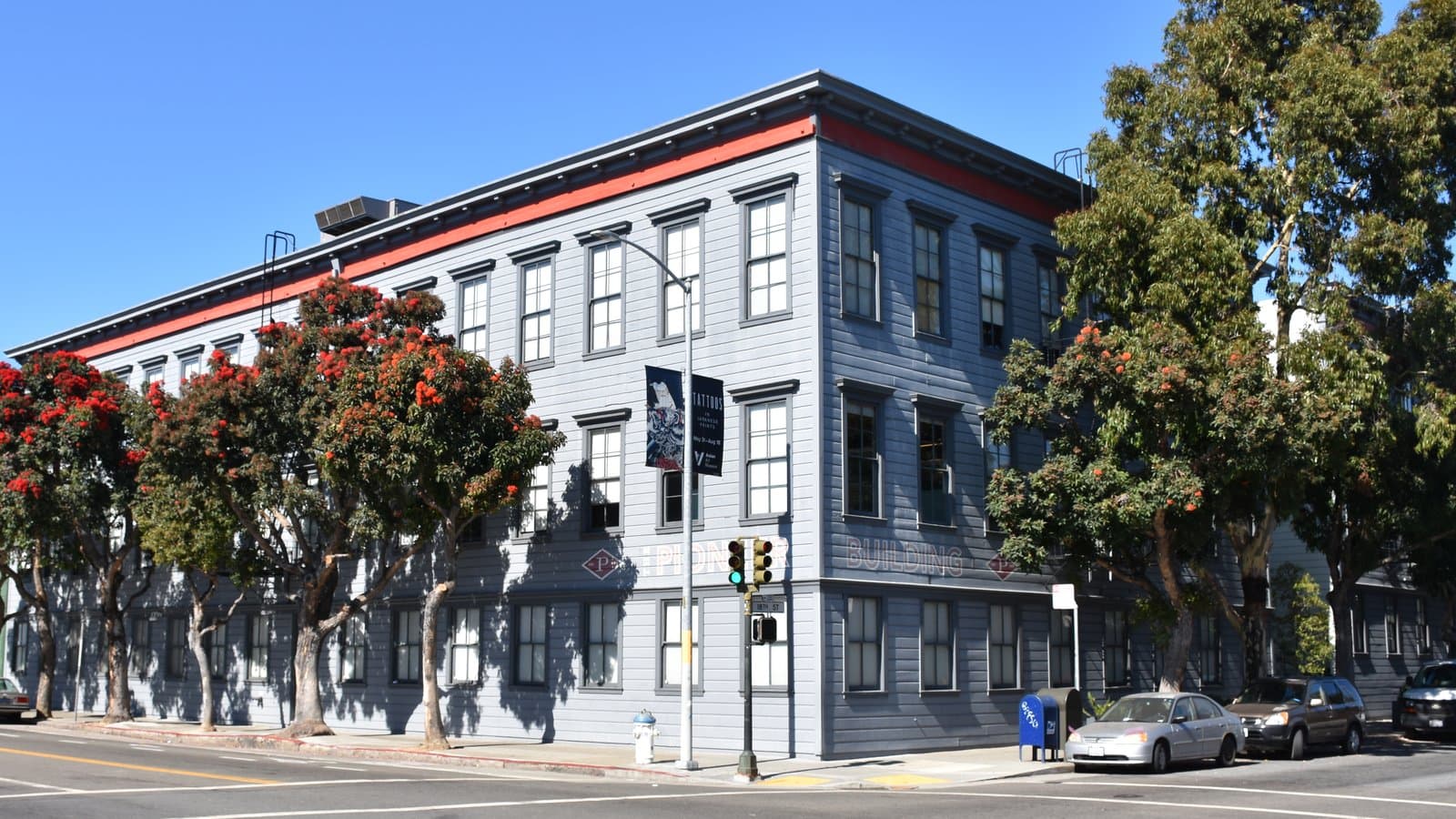 The Pioneer Building in San Francisco's Mission District, a three-story gray building with red trim on a street corner lined with flowering trees.