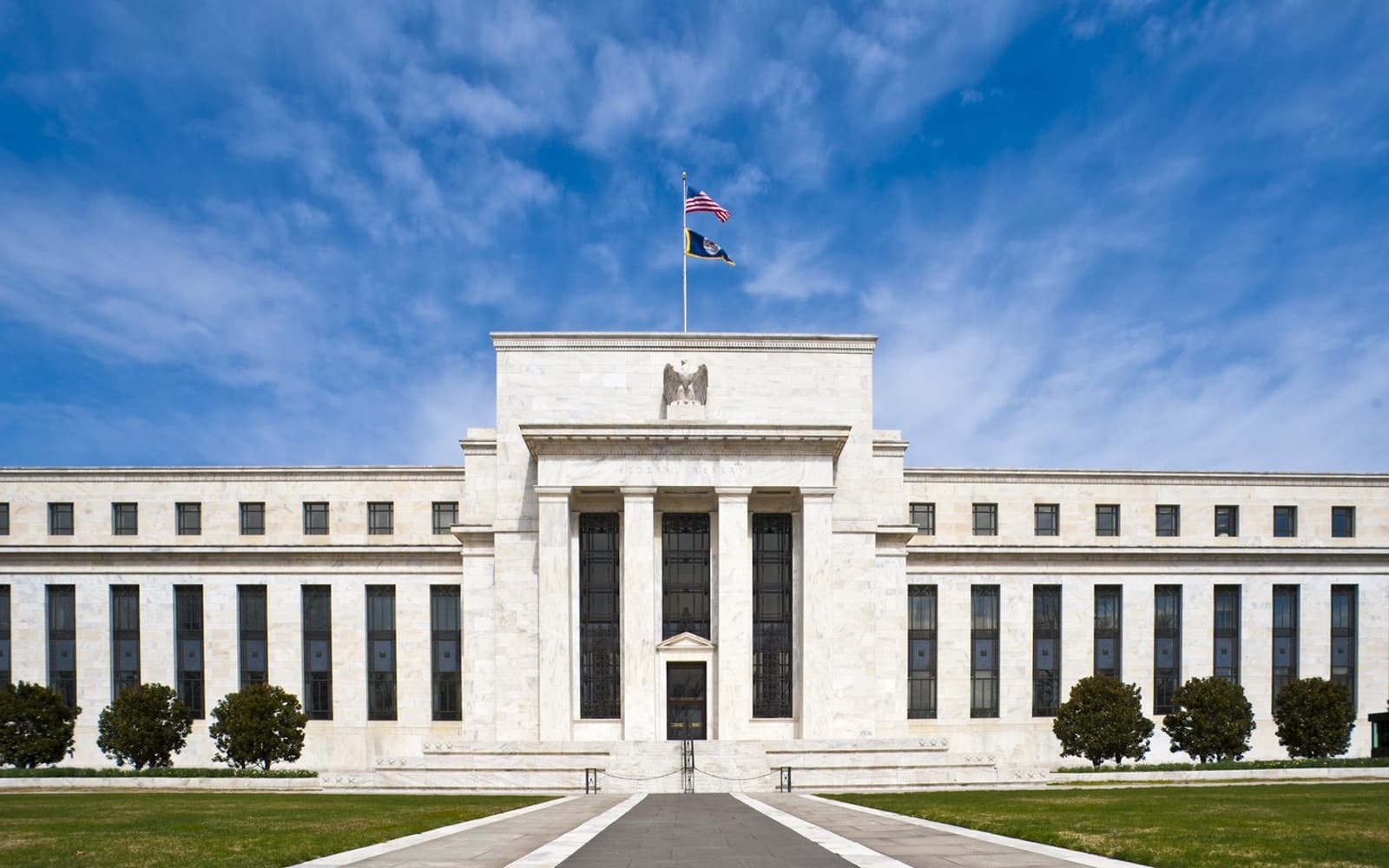 La fachada de mármol blanco del edificio Eccles de la Reserva Federal en Washington, con columnas en la entrada central y una bandera de Estados Unidos ondeando en lo alto bajo un cielo azul.